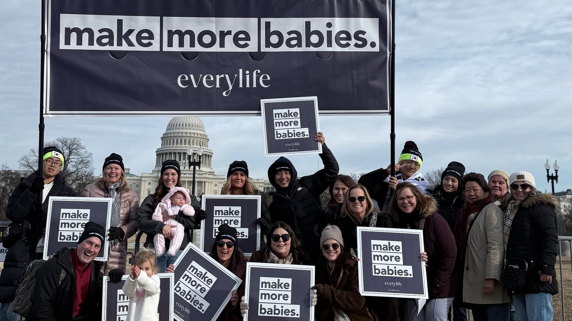 Group of people holding signs with a prominent 'make more babies' sign in front of a building.